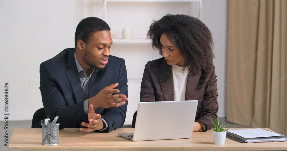 Two colleagues businesspersons man and woman co-workers sitting at table in modern office looking at laptop discussing work budget male insurance agent helps girl with finances consults using computer