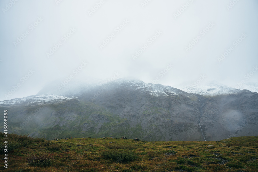 Minimalist alpine landscape with snow-capped mountains in overcast weather in rain. Deep gorge on background of snowbound mountain range under low cloudy sky. Atmospheric minimalism in high mountains.