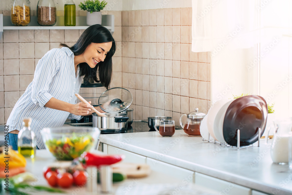 Beautiful happy young woman is cooking in the home kitchen and testing some soup from the pan on the stove Beautiful happy young woman is cooking in the home kitchen and testing some soup from the pan on the stove