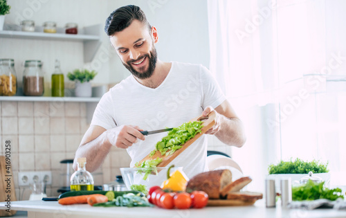 Foto Handsome happy bearded man is preparing wonderful fresh vegan salad in the kitch