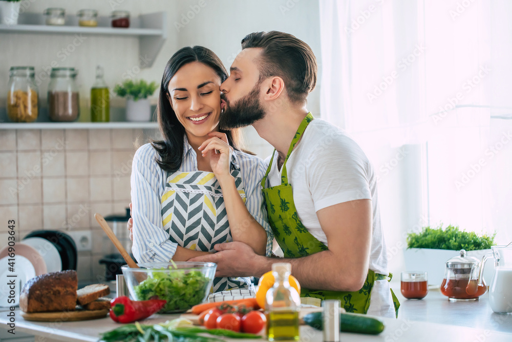Excited smiling young couple in love making a super healthy vegan salad with many vegetables in the kitchen and having fun