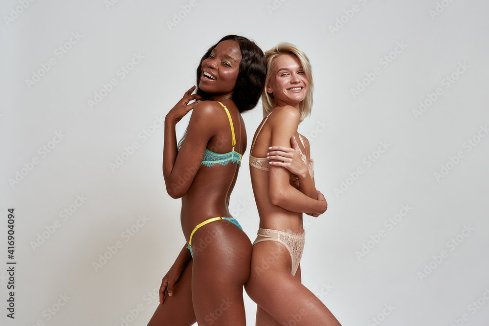 Foto de Happy african american and caucasian young women in sexy lingerie  smiling at camera, standing back to back, having fun while posing isolated  over light background do Stock