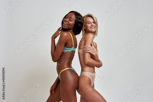 Happy african american and caucasian young women in sexy lingerie smiling at camera, standing back to back, having fun while posing isolated over light background