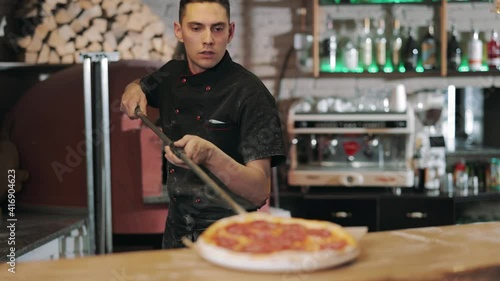 Competent chef cook in black uniform taking out of stove hot baked pizza using long shovel. Pizza maker serving tasty italian meal on round plate at restaurant. photo