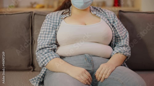 Confused overweight woman in protective mask touching fat around waist, diet photo
