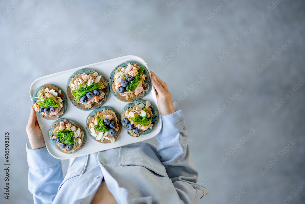 Woman in blue shirt holding a set of american pancakes on a plate with seaweed, cheese, blueberry and hummus. Homemade tasty appetizer. Top view. Copy space.