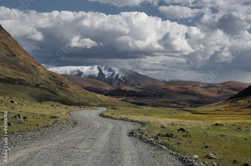 Road through a dry desert steppe on a highland mountain plateau with yellow green grass with ranges of  snow peaks rocks on a horizon skyline