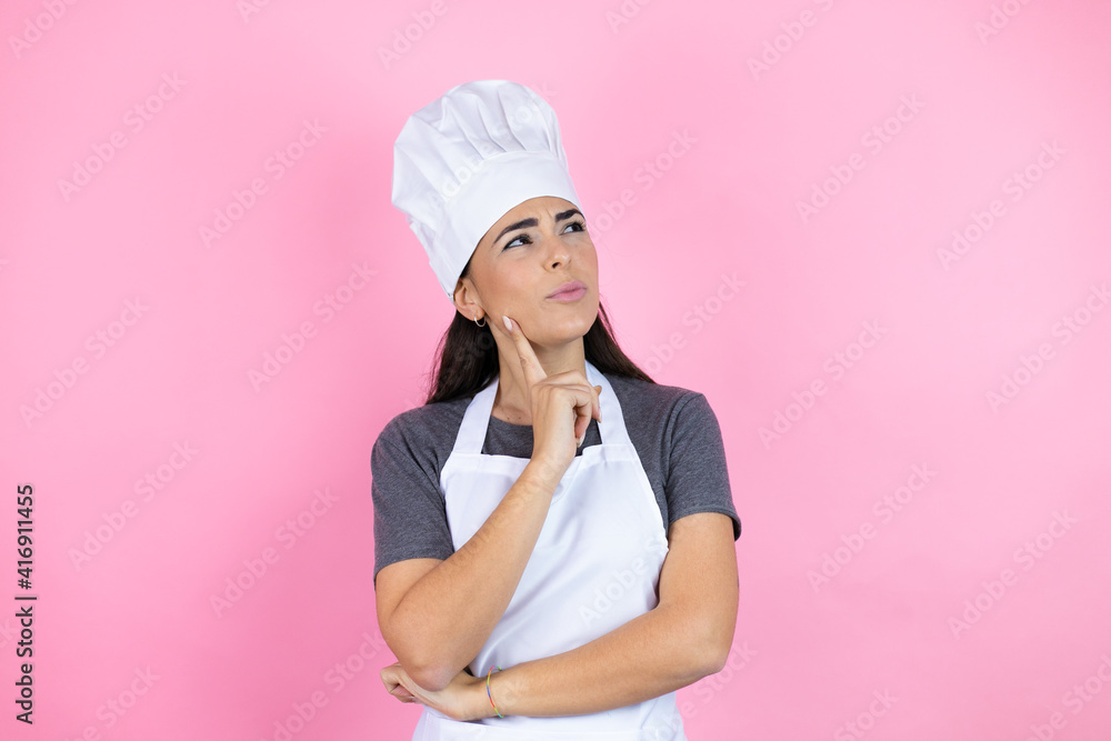 Young hispanic woman wearing baker uniform over pink background serious face thinking about question with hand on chin, thoughtful about confusing idea Young hispanic woman wearing baker uniform over pink background serious face thinking about question with hand on chin, thoughtful about confusing idea