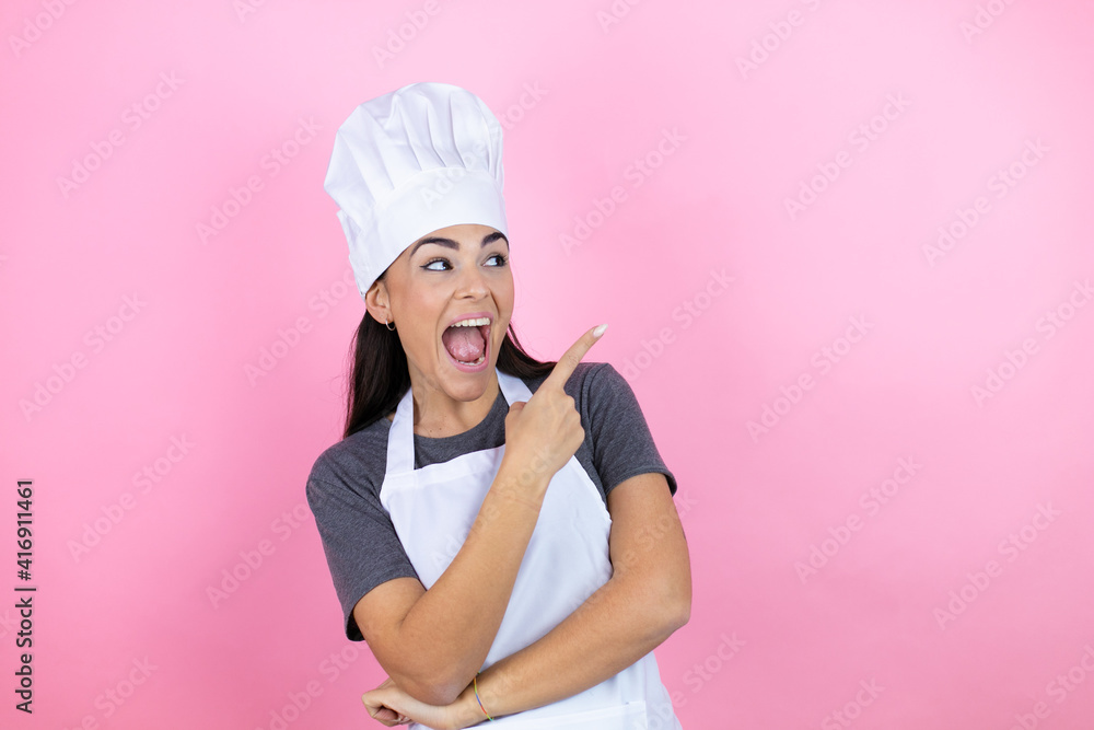 Young hispanic woman wearing baker uniform over pink background amazed and pointing with hand and finger to the side Young hispanic woman wearing baker uniform over pink background amazed and pointing with hand and finger to the side