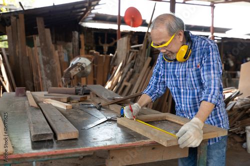 senior asian man carpenter wearing glasses and headphone, measuring wood with tape measure in workshop