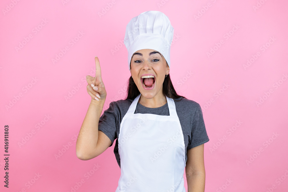 Young hispanic woman wearing baker uniform over pink background pointing finger up with successful idea. Exited and happy. Number one. Young hispanic woman wearing baker uniform over pink background pointing finger up with successful idea. Exited and happy. Number one.