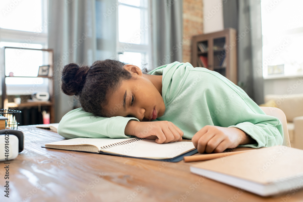 Tired mixed-race schoolgirl in hoodie keeping head on open copybook on table Tired mixed-race schoolgirl in hoodie keeping head on open copybook on table