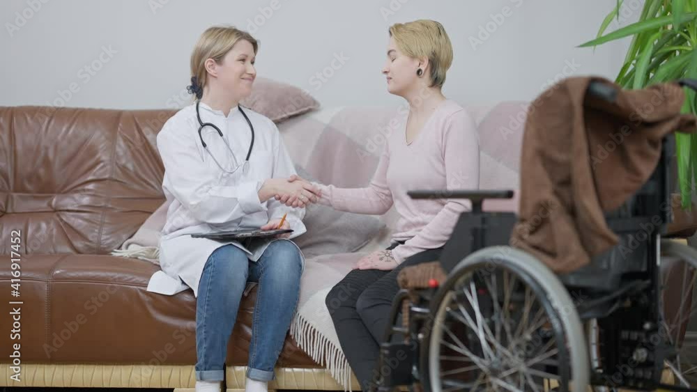 Wide shot portrait of smiling Caucasian doctor shaking hands with paraplegic patient sitting on couch indoors, and leaving. Professional specialist consulting ill paralyzed woman at home. Wide shot portrait of smiling Caucasian doctor shaking hands with paraplegic patient sitting on couch indoors, and leaving. Professional specialist consulting ill paralyzed woman at home.