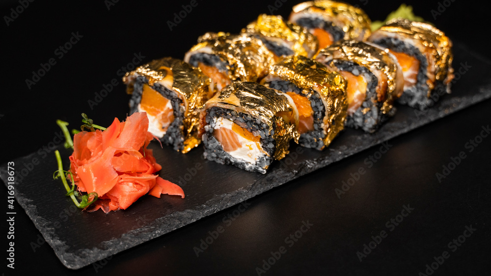 Custom sushi roll with nori, cuttlefish ink, fresh salmon, cream cheese, pepper pumpkin, eel, unagi sauce and edible gold leaf with red ginger, wasabi on a square plate on black table and background. Custom sushi roll with nori, cuttlefish ink, fresh salmon, cream cheese, pepper pumpkin, eel, unagi sauce and edible gold leaf with red ginger, wasabi on a square plate on black table and background.