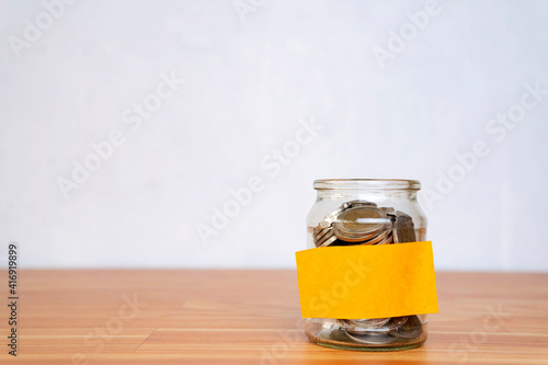 Glass jar with coins, with a label on the jar on a wooden table, white isolated background.