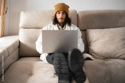 Hombre joven con el pelo largo y un gorro, cazadora vaquera, camisa, pantalón negro y botas, usando su ordenador portátil gris de metal
