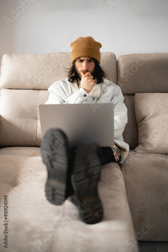 Hombre joven con el pelo largo y un gorro, cazadora vaquera, camisa, pantalón negro y botas, usando su ordenador portátil gris de metal pensando
