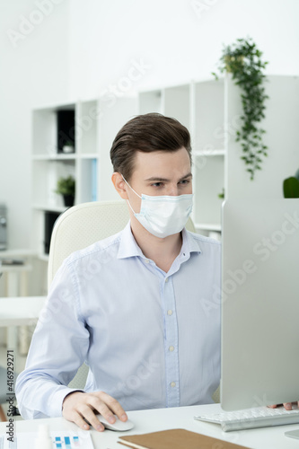Young serious designer with protective mask on face looking at computer screen