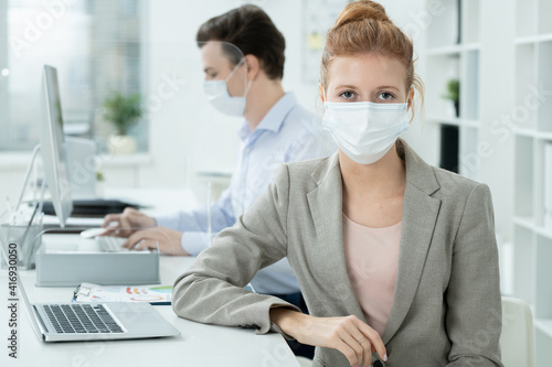 Young elegant businesswoman in protective mask looking at you against colleague