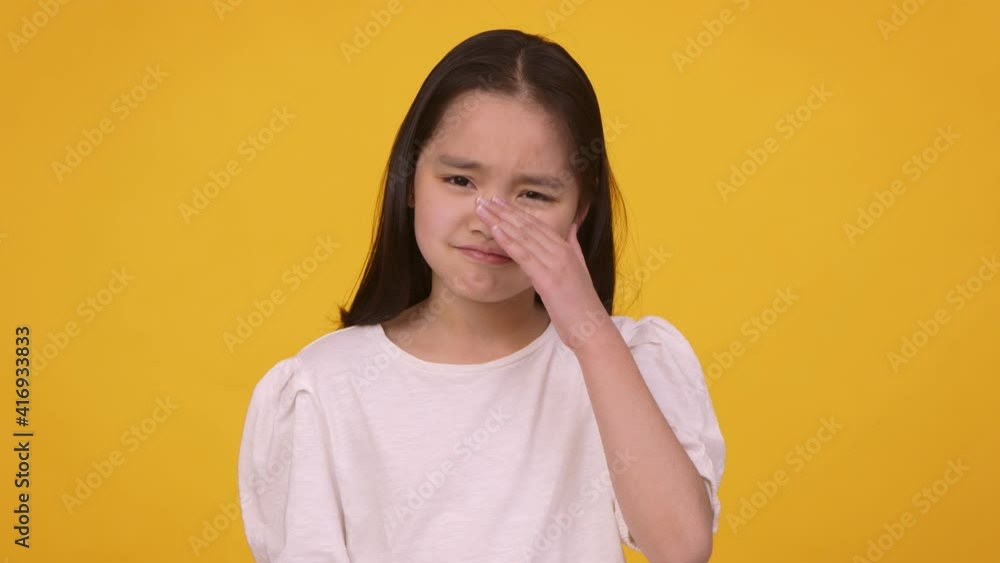 Kids problems. Portrait of upset asian girl crying and wiping her tears, orange background, slow motion Kids problems. Portrait of upset asian girl crying and wiping her tears, orange background, slow motion