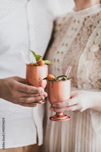 Hands of loving young couple clinking brass goblets with ice cocktail with citrus and eco-friendly glass drinking straws together, selective focus. Wedding anniversary, Valentines Day concept