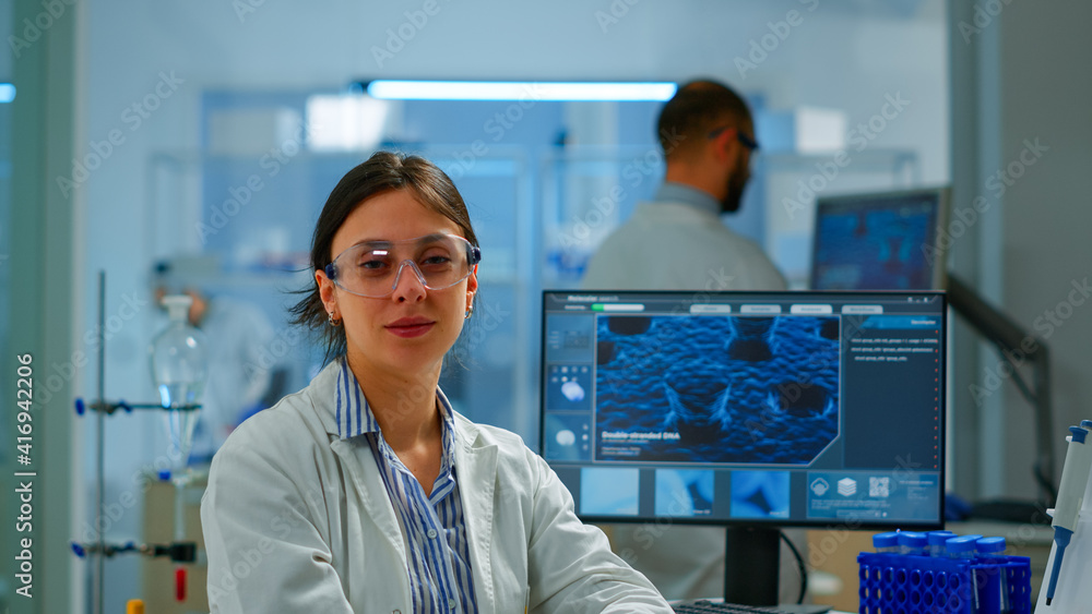 Chemist with lab coat sitting in laboratory looking at camera smiling, while colleague working on computer in background. Scientist examining virus evolution using high tech for scientific research Chemist with lab coat sitting in laboratory looking at camera smiling, while colleague working on computer in background. Scientist examining virus evolution using high tech for scientific research