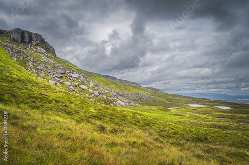 Green, long grass, large boulders and rubble on Cuilcagh Mountain mountainside with small lakes in valley below, Northern Ireland