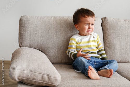 Portrait of adorable little boy sitting on the textile couch and crying. Upset toddler throwing a tantrum at home. Barefoot kid calling for attention. Close up, copy space, background.