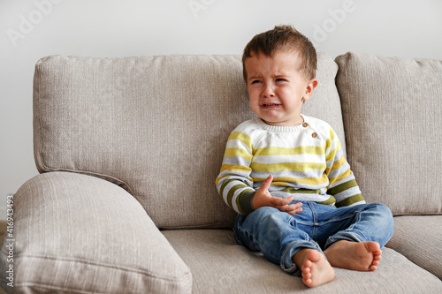 Portrait of adorable little boy sitting on the textile couch and crying. Upset toddler throwing a tantrum at home. Barefoot kid calling for attention. Close up, copy space, background.