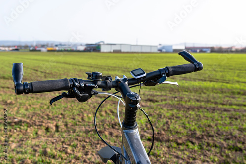 A bicycle handlebar seen from the first person perspective. Visible bicycle frame and bicycle accessories on the handlebar and the field in the background..
