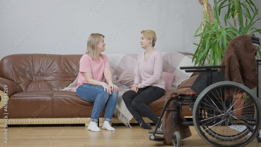 Wide shot of two cheerful young Caucasian women sitting on couch talking and laughing with blurred wheelchair at front. Positive female friends gossiping at home. Full life and paralysis. Wide shot of two cheerful young Caucasian women sitting on couch talking and laughing with blurred wheelchair at front. Positive female friends gossiping at home. Full life and paralysis.