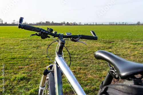 A bicycle handlebar seen from the first person perspective. Visible bicycle frame and bicycle accessories on the handlebar and the field in the background.