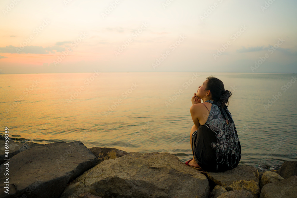 young happy and relax Asian woman enjoying sunset from beach sitting on sea shore delighted by orange sky in Summer holidays and travel concept