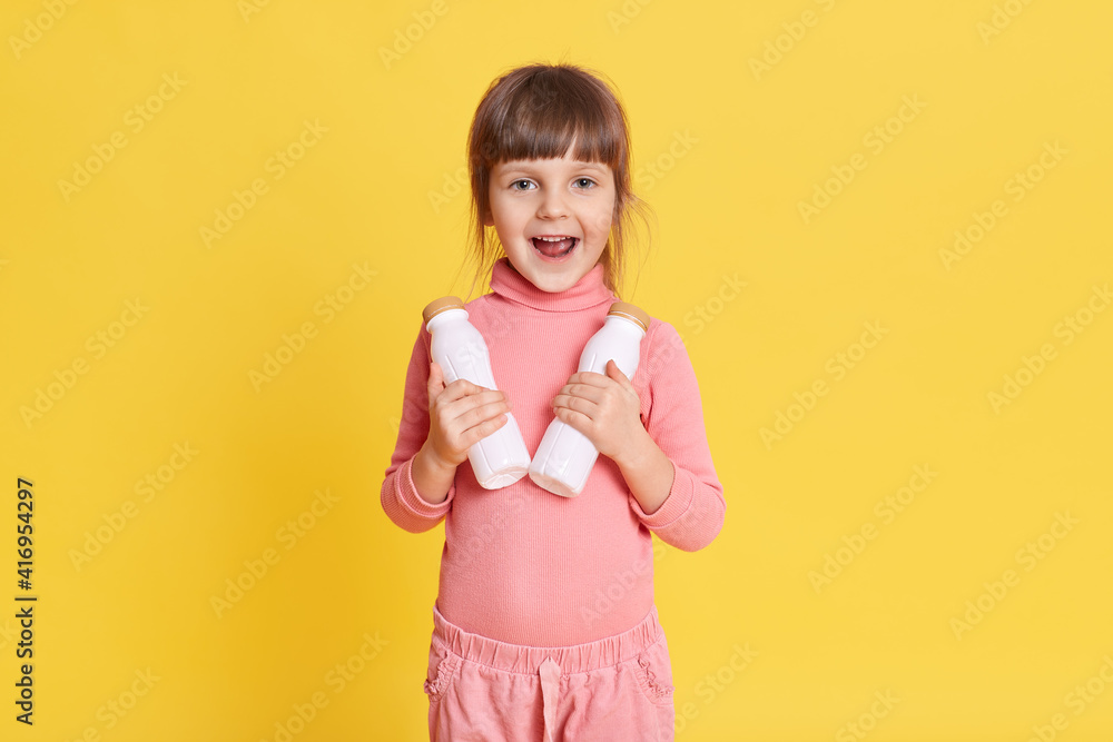 Little beauty girl in pink clothing with brown hair holding two bottles of milk and looking at camera with exited facial expression, keeps mouth opened, kid prefers healthy eating. Little beauty girl in pink clothing with brown hair holding two bottles of milk and looking at camera with exited facial expression, keeps mouth opened, kid prefers healthy eating.