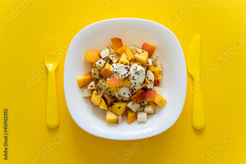 Healthy nutrition fruit salad with soft cheese and flax seeds in white plate on yellow background with yellow knife and fork. Dieting and balance food.