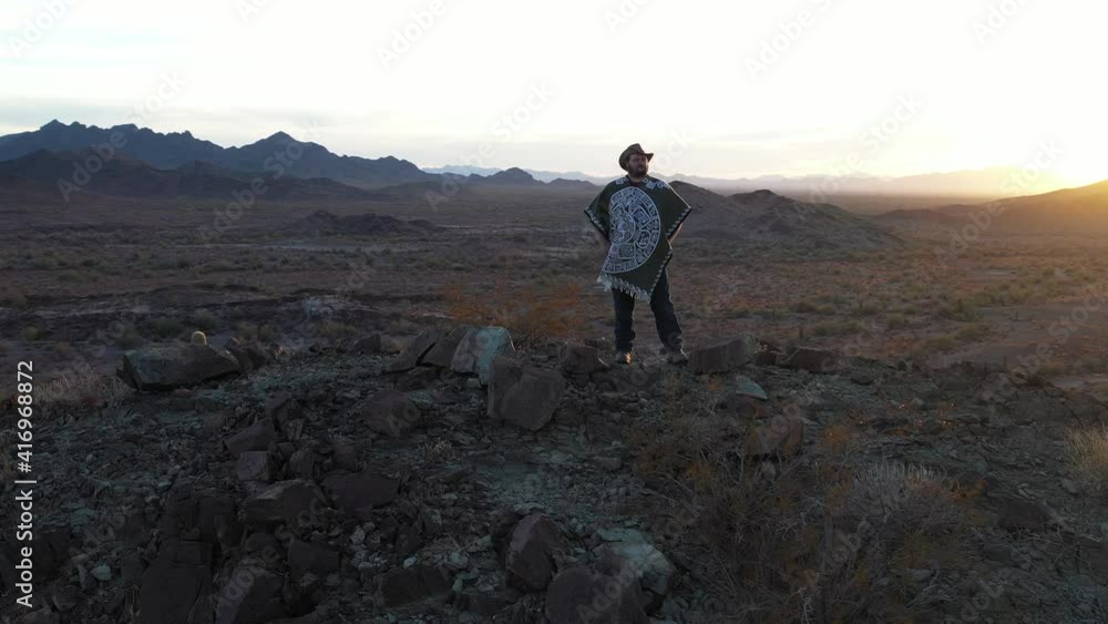 A lone man dressed as a spaghetti western hero stands atop a rugged mountain peak amidst a southwestern desert landscape as the sun sets in the distance. The camera makes a slow orbit around the man. A lone man dressed as a spaghetti western hero stands atop a rugged mountain peak amidst a southwestern desert landscape as the sun sets in the distance. The camera makes a slow orbit around the man.