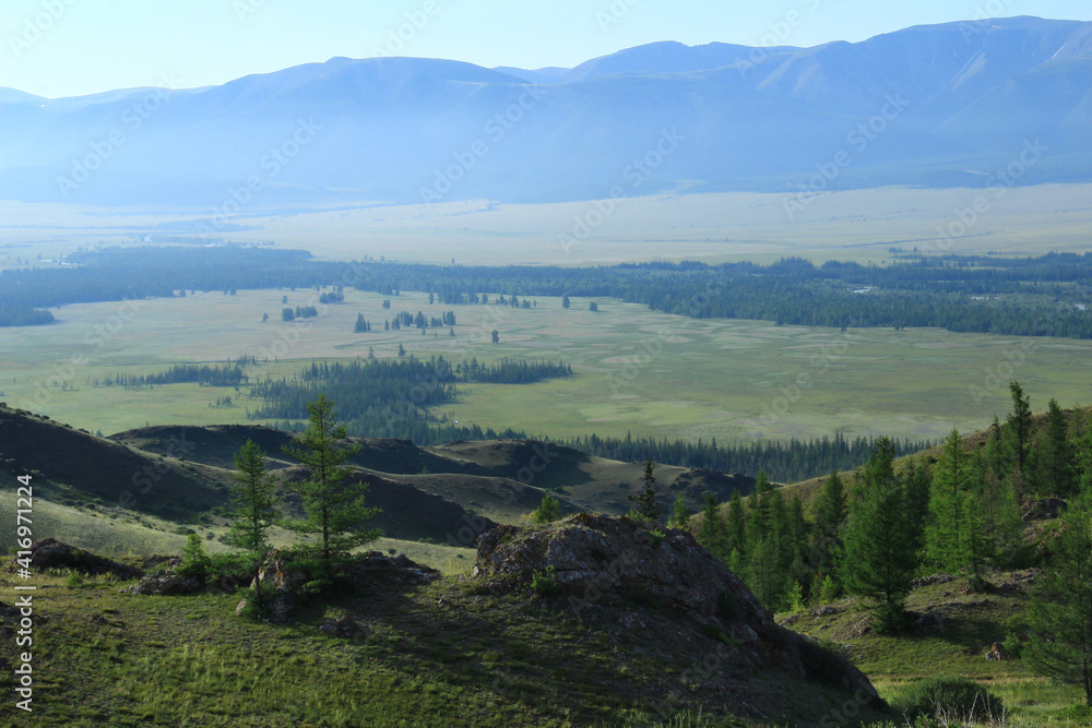 A wide alpine valley in the Kurai steppe in Altai, in the foreground a slope with rocks and trees, in the background a river, a forest and the North Chuisky ridge in the morning fog in summer