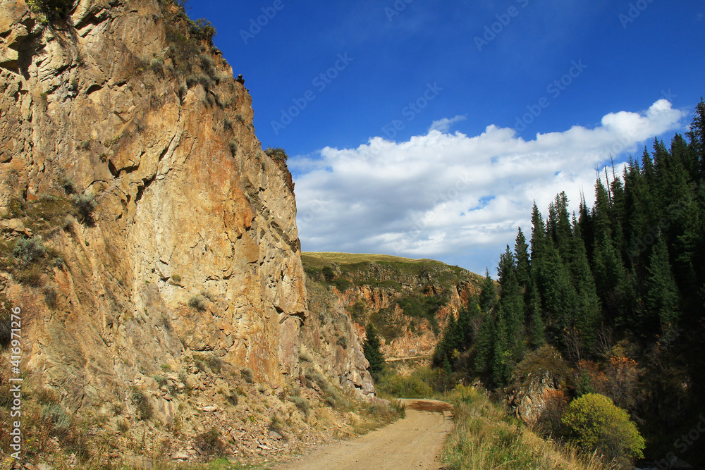 Mountain road to the high-mountainous plateau of Assy, on the right there are huge steep yellow rocks, on the left there are fir trees, the sky with a beautiful cloud, summer, sunny Mountain road to the high-mountainous plateau of Assy, on the right there are huge steep yellow rocks, on the left there are fir trees, the sky with a beautiful cloud, summer, sunny