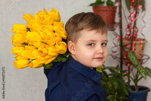 A young, beautiful boy. He carries a huge armful of fresh yellow tulips on his shoulder. The concept of spring and holiday, March 8, International Women's Day,
