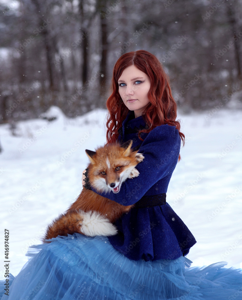 Beautiful red haired girl in blue dress with red fox in winter forest Beautiful red haired girl in blue dress with red fox in winter forest