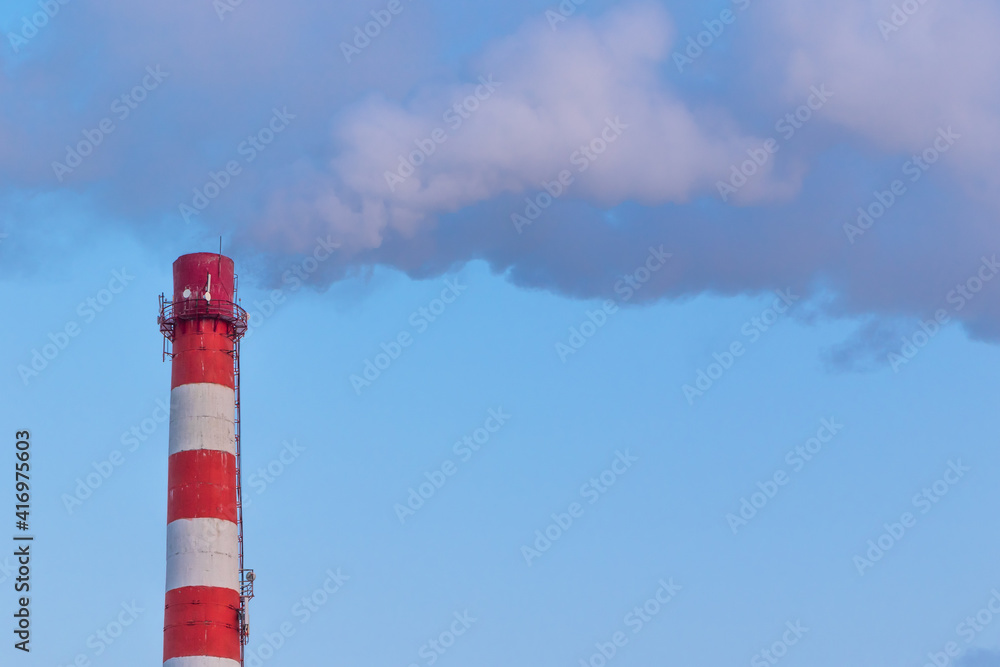 red and white chimney of a factory, from which gray smoke comes out against a blue sky on a sunny frosty winter day