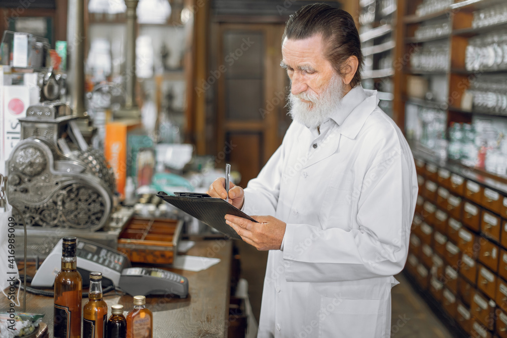 Portrait of concentrated experienced male senior pharmacist writing on a clipboard while standing in beautiful vintage old pharmacy interior. Portrait of concentrated experienced male senior pharmacist writing on a clipboard while standing in beautiful vintage old pharmacy interior.