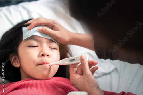 Asian mother measuring temperature girl and hand on forehead on bed at morning time, Sick child have cool gel for reduce high fever, Selective focus, Healthy and infection concept
