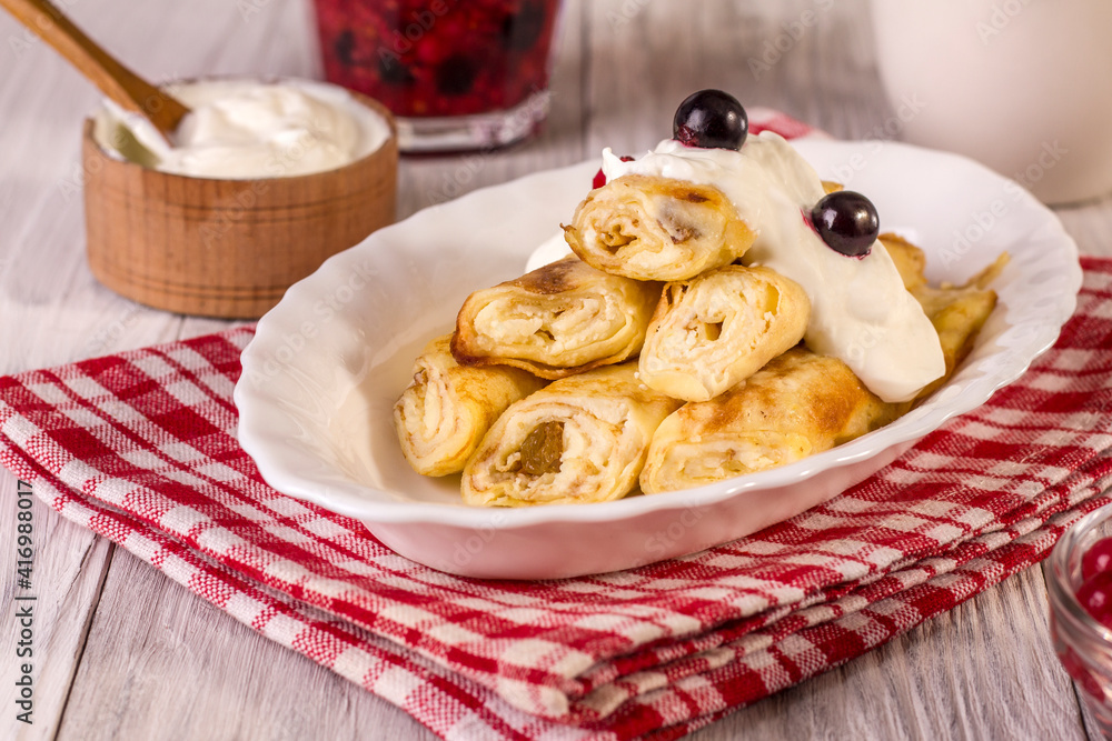 Rolled up thin pancakes stuffed with cottage cheese with berries and sour cream on a white wooden table.