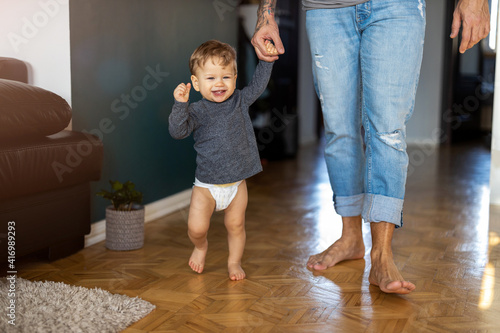 Little boy learning to walk with his father next to him at home 
