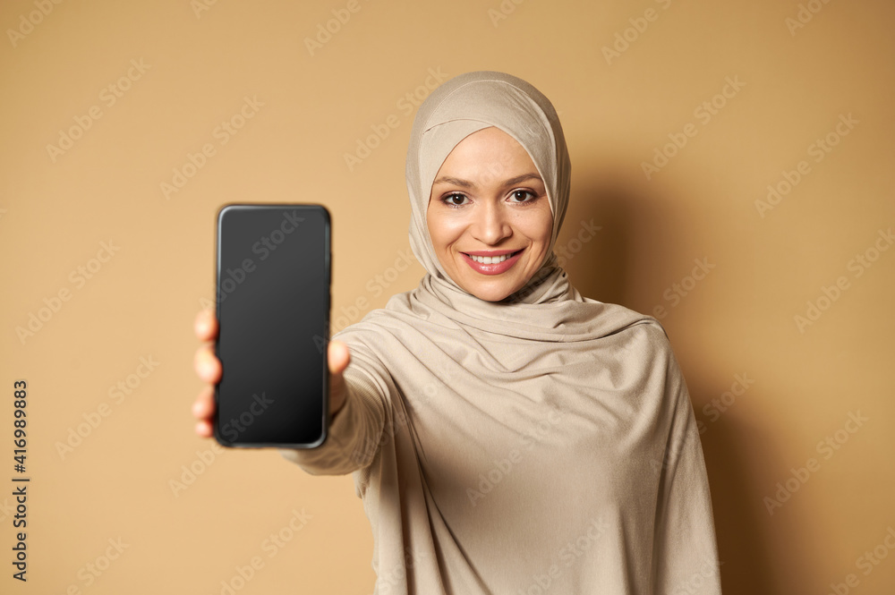 Muslim woman with covered head in headscarf smiles with toothy smile and shows a cell phone to the camera standing on beige background with copy space Muslim woman with covered head in headscarf smiles with toothy smile and shows a cell phone to the camera standing on beige background with copy space