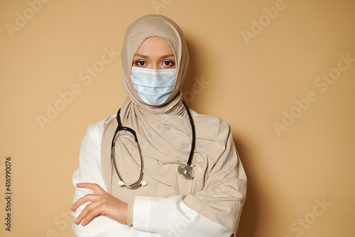 Muslim woman doctor in hijab and medical mask looking at camera with crossed arms. Beige background with copy space photo