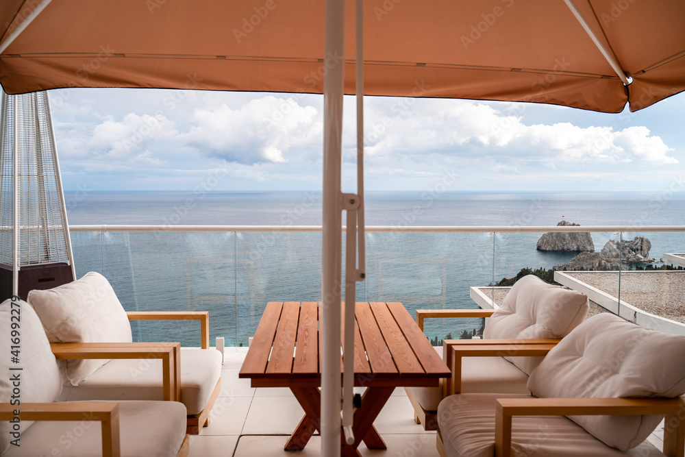 Restaurant with sea view. You can see rocks in the sea. Wooden tables and chairs with white cushions. Overcast, cloudy weather.
