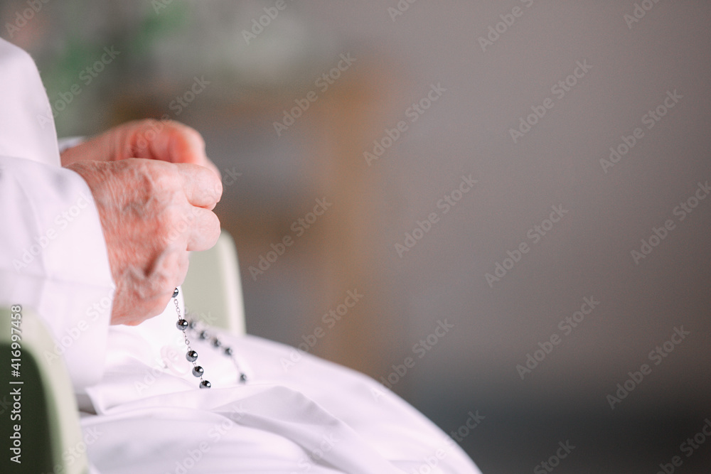 Nun dressed in white and sitting on the chair holding the rosary in hand and praying. Selective focus. Hope concept