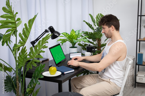 A young employee sits at a desk and types on a laptop keyboard. Remote work at home. A domain computer workstation among potted plants with bright lighting. Green screen of monitor ready to design.